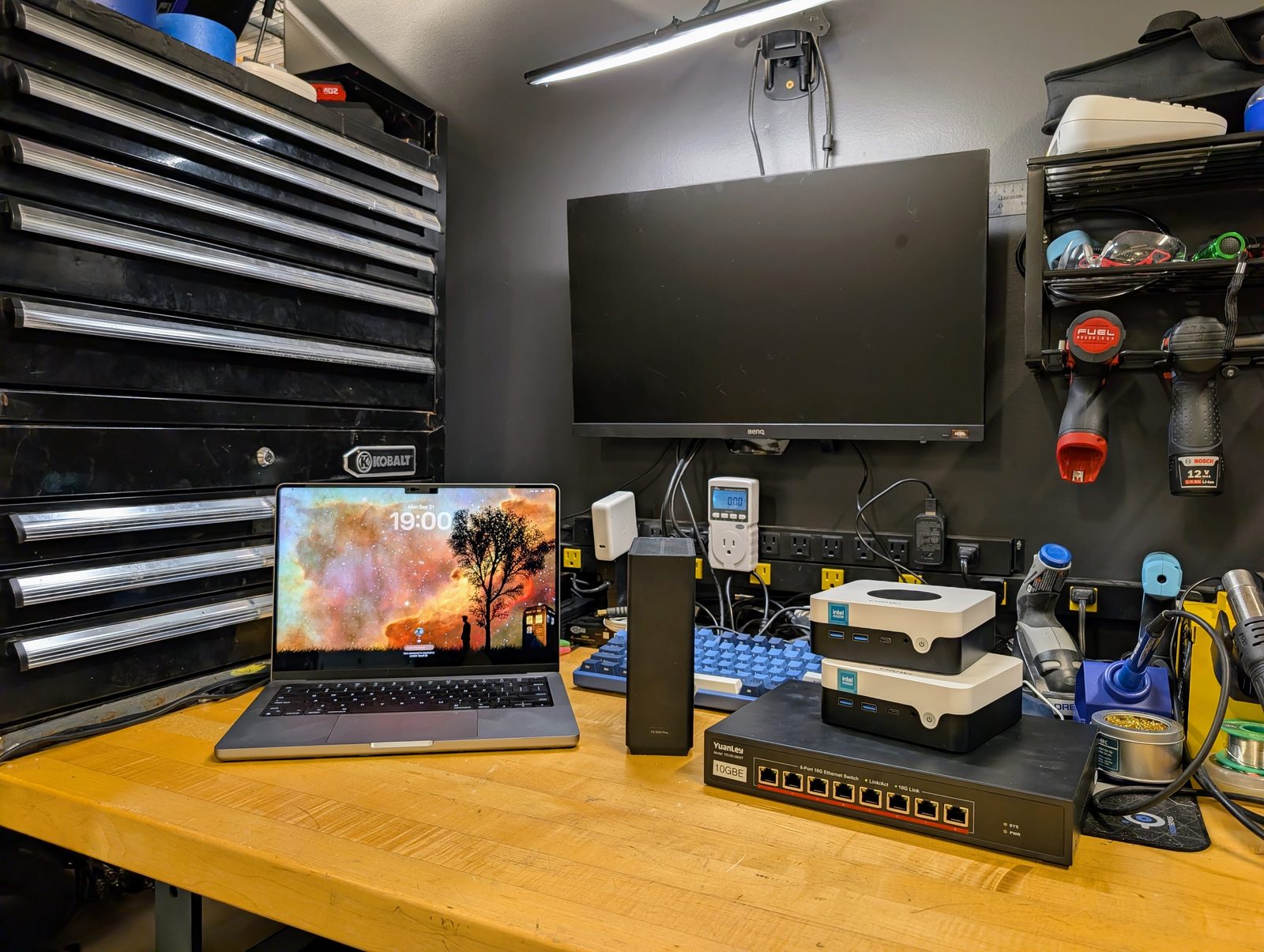 A workbench with a laptop, two small white computers, a small network switch and a monitor, tools on the wall behind