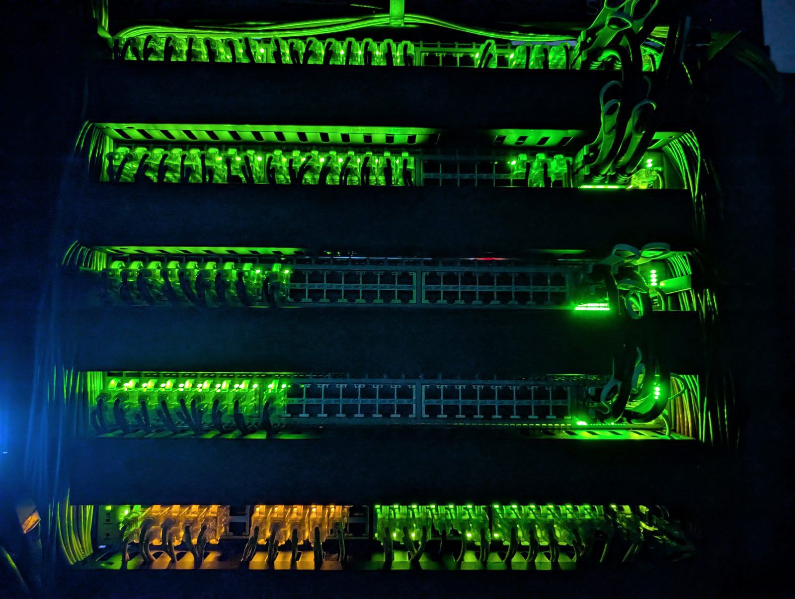 Five rows of network switches in the dark, every patched port lit green and amber