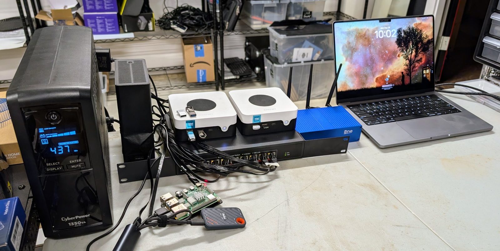 A workbench with a tower UPS showing its runtime, a small black eight-bay storage box, two small white mini PCs on a 10 GbE switch, a blue router with three antennas, a Raspberry Pi with a portable SSD, and a laptop