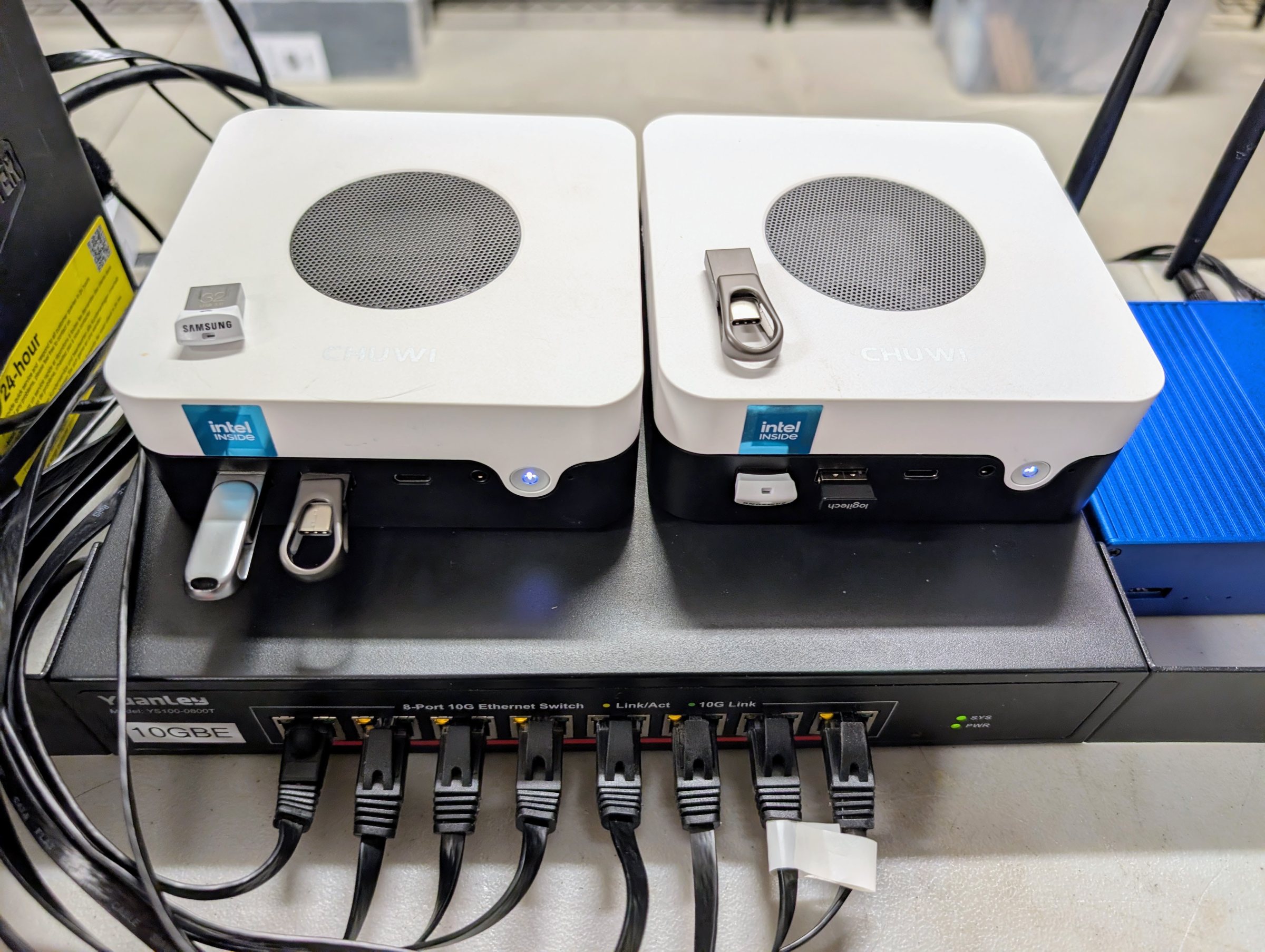 Two white mini PCs with round top vents sitting on an 8-port 10G switch, USB sticks in their front ports, and a network cable in every switch port.