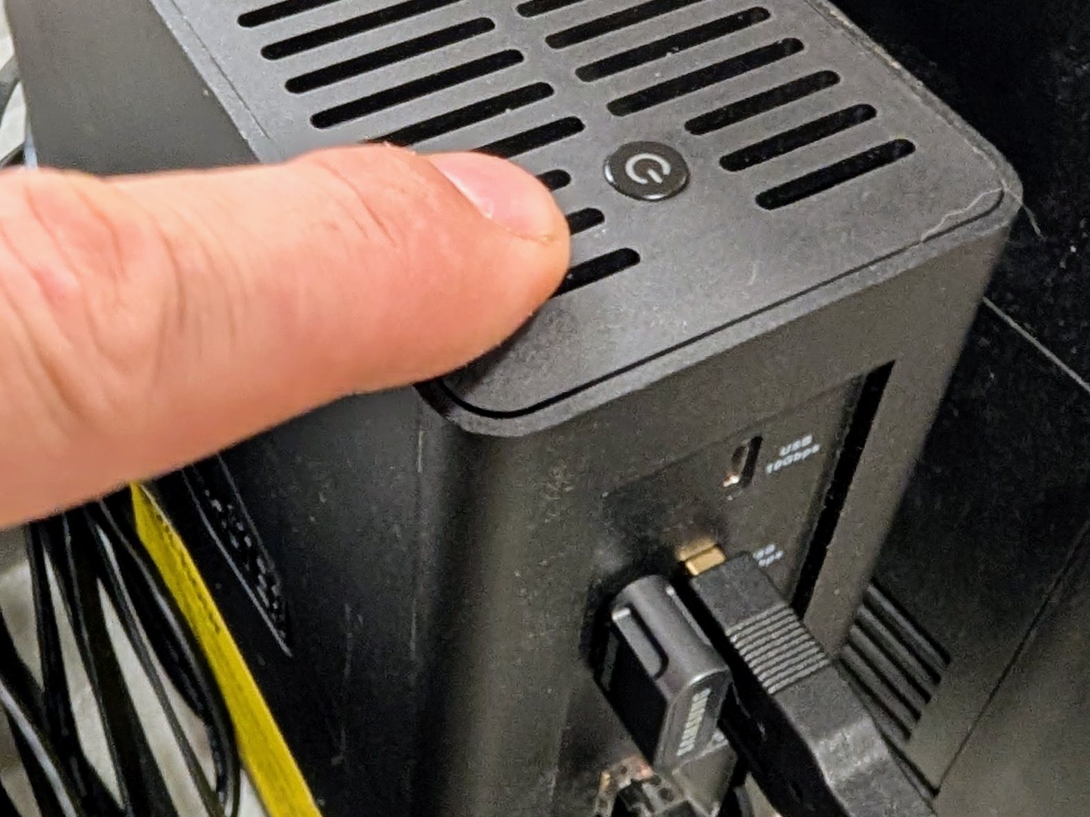 A finger resting on the top panel of a black NAS next to its round power button; directly below, on the back panel, two USB plugs are in the ports.