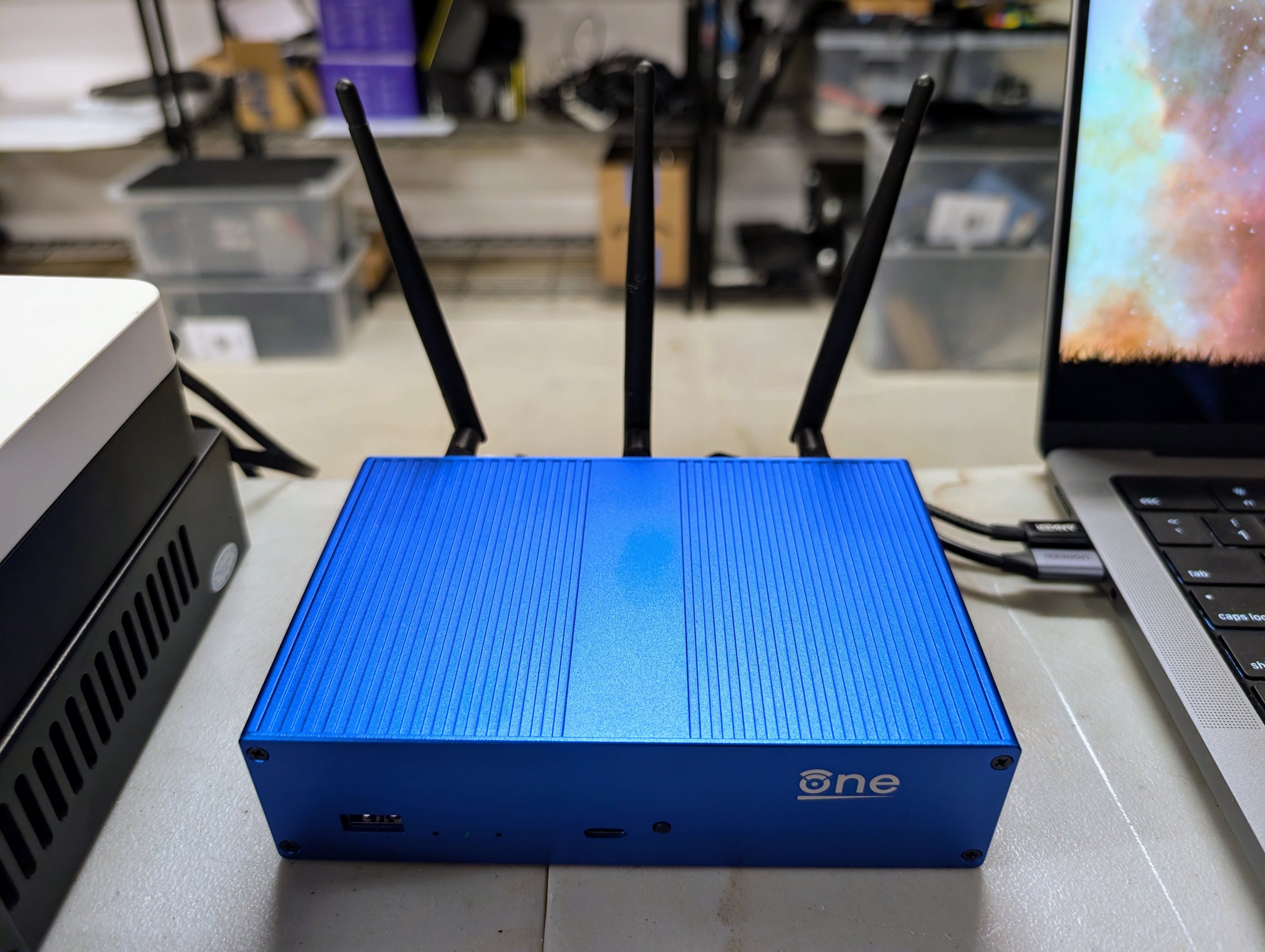 A blue metal router with three black antennas, between a mini PC and a laptop
