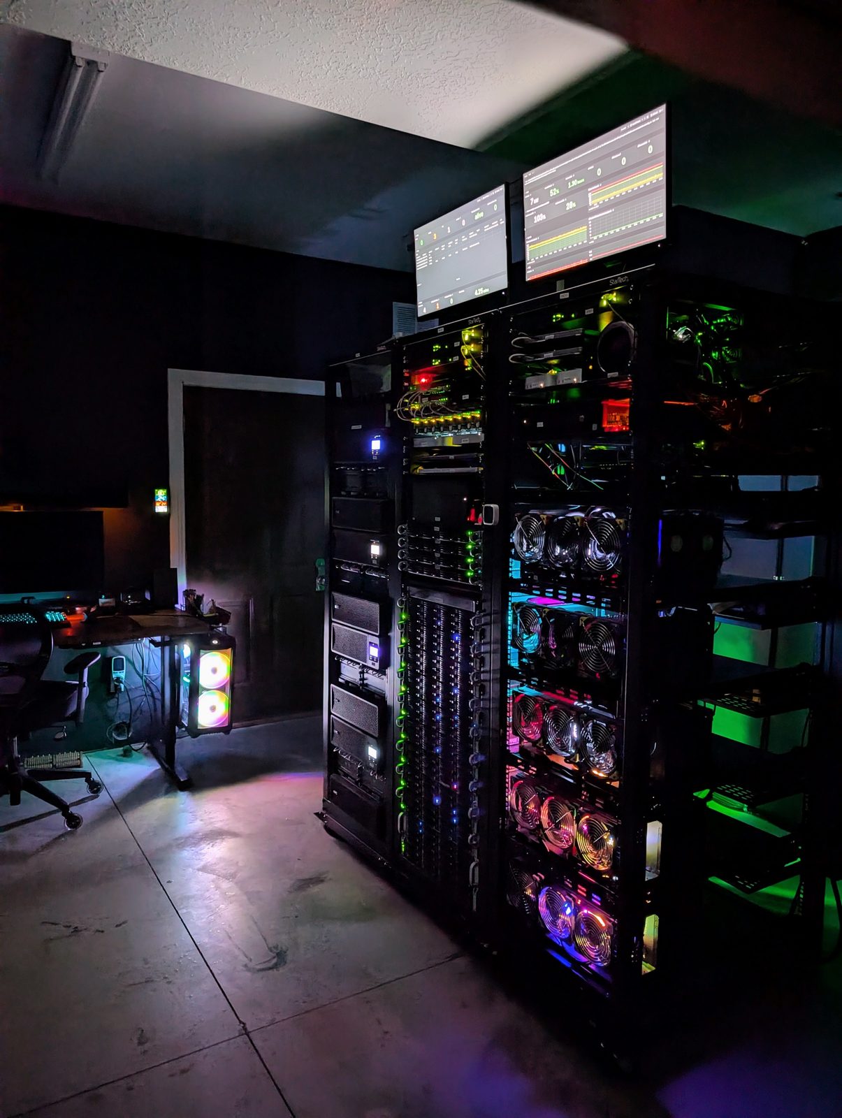 A dark studio: a desk with a glowing workstation, and beside it three server racks lit by their own LEDs and two dashboards